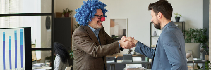 Poster - Header of happy mature male boss in clown wig and nose shaking hand of young businessman while greeting him in office