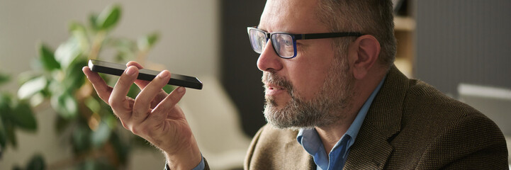 Poster - Header of mature male boss in elegant suit recording voice message while sitting in front of camera and holding smartphone by his mouth