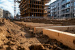 © Tondone - An urban construction site featuring wooden planks laid on a dirt foundation with a partially built building in the background under a clear blue sky.