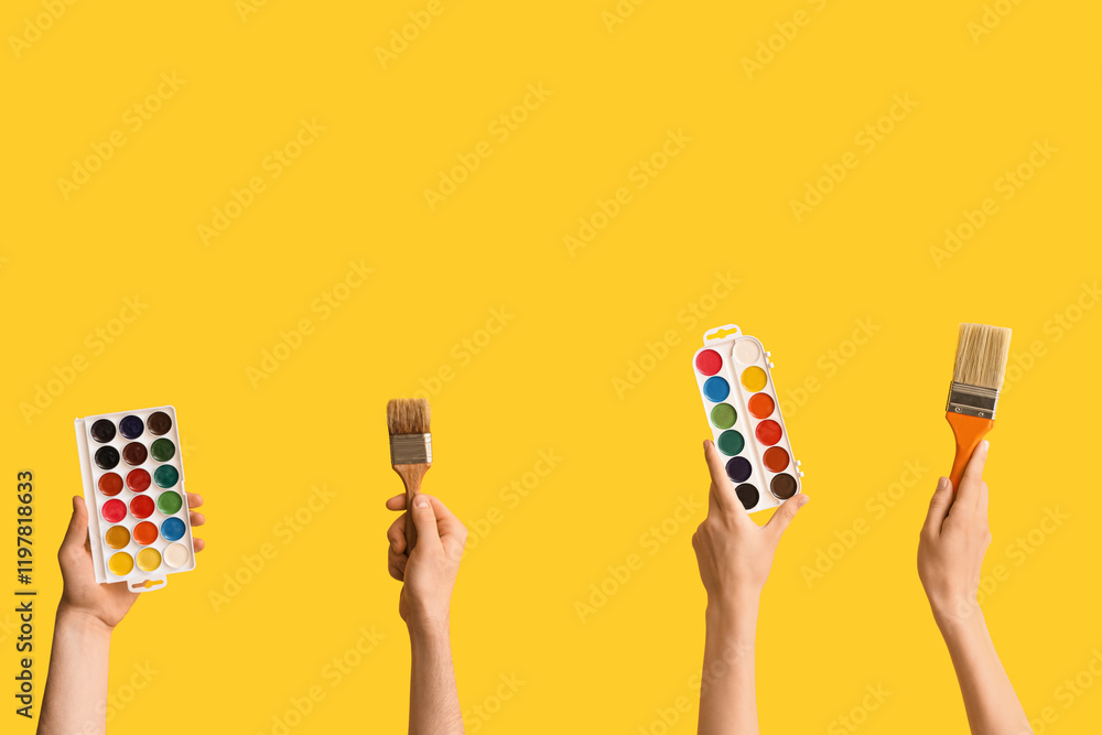 Female hands with paintbrushes and watercolor paints on yellow background