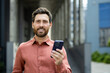 © Liubomir - Portrait of successful businessman man with phone in hands. Office worker smiling and looking at camera, using app on smartphone, walking in city from outside office building.