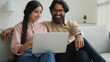 © Yuliia - Arabian Indian couple on couch sofa use computer laptop shopping online internet store happy multiracial wife husband smiling lovely customers woman and man search goods purchase on website at home