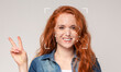 © Prostock-studio - A young woman with vibrant red hair smiles widely while making a peace sign with her hand. Facial recognition markers are displayed on her face, highlighting the technology in action.