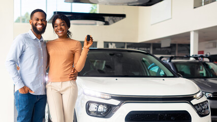 Wall Mural - Happy African American Spouses Posing Near Luxury Car In Showroom, Cheerful Black Couple Holding Keys And Smiling At Camera, Buying New Vehicle In Modern Auto Salon, Full Length Shot, Copy Space