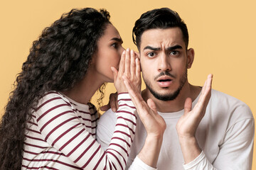 Wall Mural - Young Arab Woman Sharing Gossips With Her Shocked Boyfriend, Whispering To His Ear, Millennial Middle Eastern Female Telling Secret To Spouse While They Standing Together Over Yellow Background