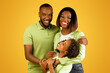 © Prostock-studio - Happy family. Loving african american parents with daughter embracing and smiling at camera over yellow background, free space, studio shot. Parenthood and bonding concept