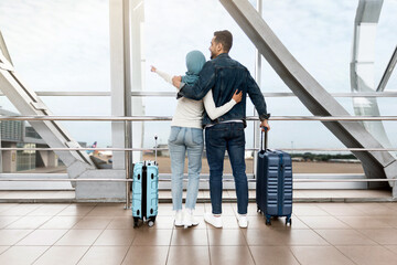 Wall Mural - Weekend Travel. Loving Islamic Couple Standing Near Window In Airport And Pointing Away, Rear View Of Muslim Spouses With Suitcases Waiting For Flight At Terminal, Enjoying Upcoming Trip, Copy Space