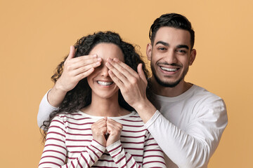 Wall Mural - Cheerful Arab Guy Playing Guess Who With His Excited Girlfriend, Covering Her Eyes With Hands And Making Surprise While Posing Together Over Yellow Studio Background, Closeup Shot With Free Space