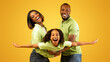 © Prostock-studio - Family bonding concept. Joyful black man and woman holding daughter on hands, having fun over yellow studio wall. Smiling girl pretending she is flying, spreading hands, imitating plane