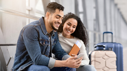 Wall Mural - Waiting For Flight. Young Arab Spouses Using Smartphone In Airport Terminal, Middle Eastern Couple Browsing Internet On Mobile Phone Or Booking Hotel Online Before Flight Boarding, Closeup Shot