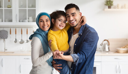 Wall Mural - Muslim Family Portrait. Happy Middle Eastern Father, Mother And Little Daughter Posing Together In Kitchen Interior, Loving Arab Parents And Cute Female Child Embracing And Smiling At Camera