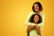 © Prostock-studio - Mother's love. Happy loving african american mom cuddling her cute daughter from the back, standing over yellow background with free space and smiling to camera