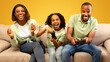 © Prostock-studio - Domestic entertainment. Emotional black dad and daughter holding joysticks, competing with each other, parents playing video games with child while sitting on sofa over yellow background