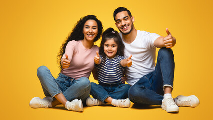 Wall Mural - Like Concept. Happy Middle Eastern Family Of Three Showing Thumbs Up At Camera While Sitting Over Yellow Studio Background, Happy Arab Parents And Little Daughter Approving Or Recommending Something