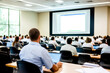 © F-STUDIO - A group of professionals attentively watching a presentation in a large conference room, with a focus on the speaker and screen.