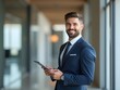 © NETI studio - Confident young professional dressed smartly, smiling and holding a tablet, standing in a modern office hallway with natural lighting, representing leadership and innovation in a business environment