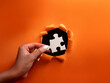 © YiuCheung - A hand places a white puzzle piece into a hole torn in bright orange paper revealing a black background.  This symbolizes problem-solving and finding solutions.