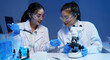 © Phimwilai - Two female scientists wearing lab coats and safety glasses are conducting research in a modern laboratory, using a pipette and microscope with blue liquid samples and test tubes
