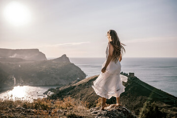 Wall Mural - A woman in a white dress stands on a hill overlooking the ocean. The scene is serene and peaceful, with the woman's dress billowing in the wind. The combination of the ocean.