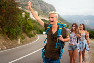 Wall Mural - cheerful interracial friends standing on road showing hitchhiking gestures. tourism traveling