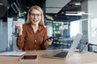 © Liubomir - Portrait of successful woman sitting at desk inside office at workplace. Mature businesswoman holding phone in hands and celebrating triumph and successful achievement results. Looking at camera.