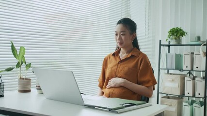 Wall Mural - Medium long shot of cheerful Asian pregnant businesswoman sitting in front of laptop while calling on video at office desk