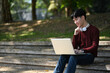 © saltdium - A young man in a sweater, headphones resting around his neck, sits on outdoor steps with a laptop, enjoying a peaceful green environment as he works or studies