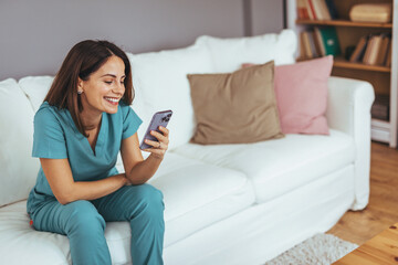 Wall Mural - Smiling Woman in Scrubs Using Smartphone on Comfortable White Sofa