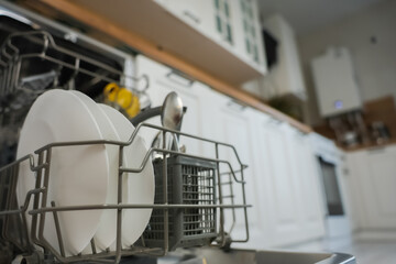 Wall Mural - Open dishwasher close-up with clean white plates and cutlery on the left of the photo against the background of a white wooden kitchen out of focus. The concept of washing dishes, dishwashers