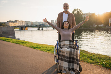 Wall Mural - Senior woman in wheelchair walking with caregiver old man on road. Family couple man husband supporting taking care of paralyzed woman wife in chair for people with disability outdoor. Rehabilitation