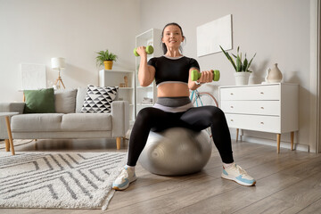 Wall Mural - Sporty young woman with dumbbells training on fitball at home