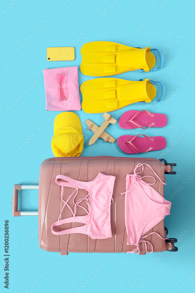 Suitcase with beach accessories and wooden airplane on blue background, top view