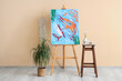 © Pixel-Shot - Easel with painting, houseplant and art supplies on stool near beige wall