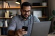 © Milos - A young man is seated in a modern workspace, engrossed in his phone and laptop, reflecting the intersection of technology, work, and daily life in a contemporary setting.