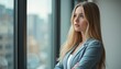 © natakot - A thoughtful young woman with long blonde hair stands by a large window, lost in contemplation. Dressed in a stylish gray blazer, she gazes at the city skyline, embodying ambition and introspection in