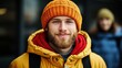 © JO BLA CO - A young man with a beard stands in an urban environment, wearing a bright orange hat and a yellow coat. He smiles warmly as daylight enhances the winter atmosphere
