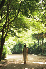 Wall Mural - A woman in a white dress walks down a sunlit path surrounded by lush, green trees, creating a serene and peaceful atmosphere.