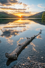 Wall Mural - A tranquil lake at sunset with a fallen log in the foreground and mountains silhouetted in the background, reflecting vibrant orange and blue hues in the water.