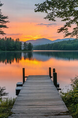 Wall Mural - A serene lake landscape during sunset with a wooden dock extending into calm, mirrored waters, surrounded by trees and mountains in the background. Glowing orange and pink sky.