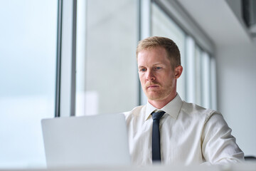 Wall Mural - Young business man executive wearing tie working on laptop in office at desk near big window. Busy businessman financial analyst or manager using computer technology at work. Close up photo.