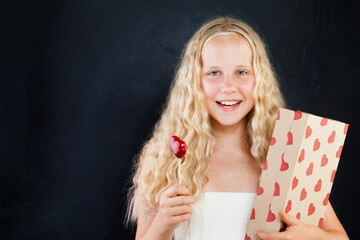 Wall Mural - Happy surprised young girl holding gift box with red heart. Blond child with long healthy curly hair on black background