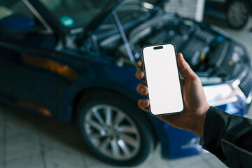 Fototapete - Hand holding a smartphone with a blank screen in front of an open car hood, showing the engine compartment. Concept for car diagnostics, automotive apps, or remote vehicle monitoring