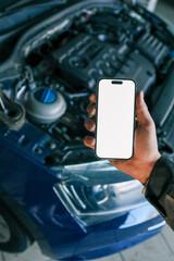 Fototapete - Hand holding a smartphone with a blank screen in front of an open car hood, showing the engine compartment. Concept for car diagnostics, automotive apps, or remote vehicle monitoring