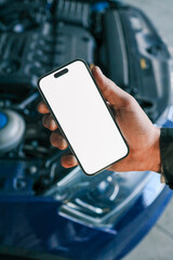Fototapete - Hand holding a smartphone with a blank screen in front of an open car hood, showing the engine compartment. Concept for car diagnostics, automotive apps, or remote vehicle monitoring