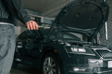 Fototapete - Hand holding an adjustable wrench in an automotive repair shop. A blue car with an open hood is visible in the background, highlighting repair or maintenance activity
