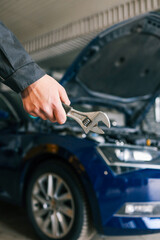 Fototapete - Hand holding an adjustable wrench in an automotive repair shop. A blue car with an open hood is visible in the background, highlighting repair or maintenance activity