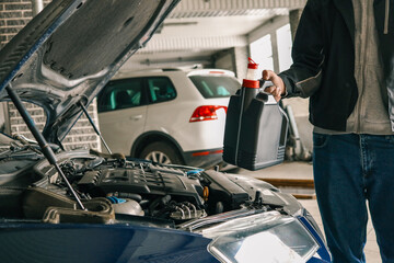 Fototapete - Mechanic holding a bottle of motor oil near the open hood of a car, ready for an oil refill in an automotive workshop. Scene depicts routine car maintenance