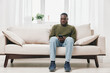 © SHOTPRIME STUDIO - Thoughtful Black man sitting on a light sofa, checking his phone, in a bright and modern living room filled with natural light and greenery