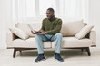 © SHOTPRIME STUDIO - Young African man working on a laptop at home, dressed in a green sweater, sitting on a light sofa, conveying a relaxed and focused emotion while surrounded by a bright, cozy atmosphere