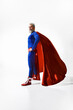 © master1305 - Gray-haired man in blue superhero outfit and red cape stands tall and determined, showing strength and resilience against white studio background. Concept of human emotions, empowerment